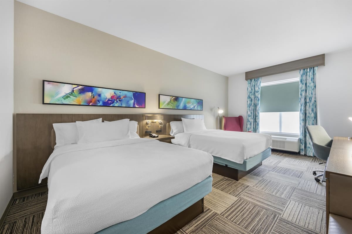 A hotel room with two beds, a desk, a chair, and artwork on the wall.