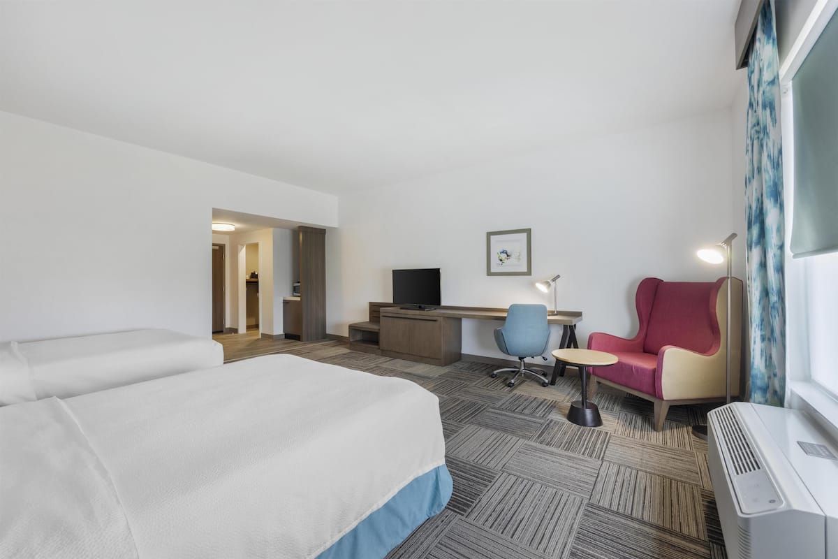 A hotel room with a large bed, a desk with a computer, a chair, a small table, and a window with curtains.