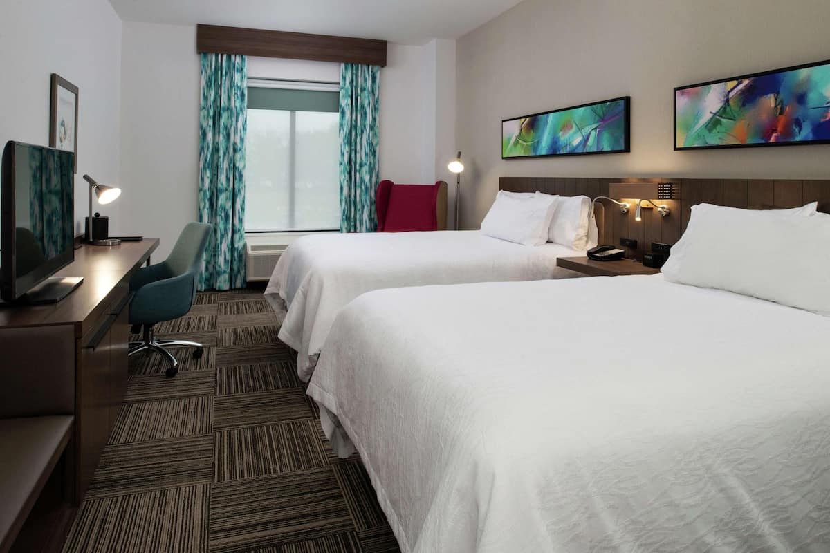 A hotel room with two beds, a desk with a computer, a chair, a TV, and artwork on the walls.