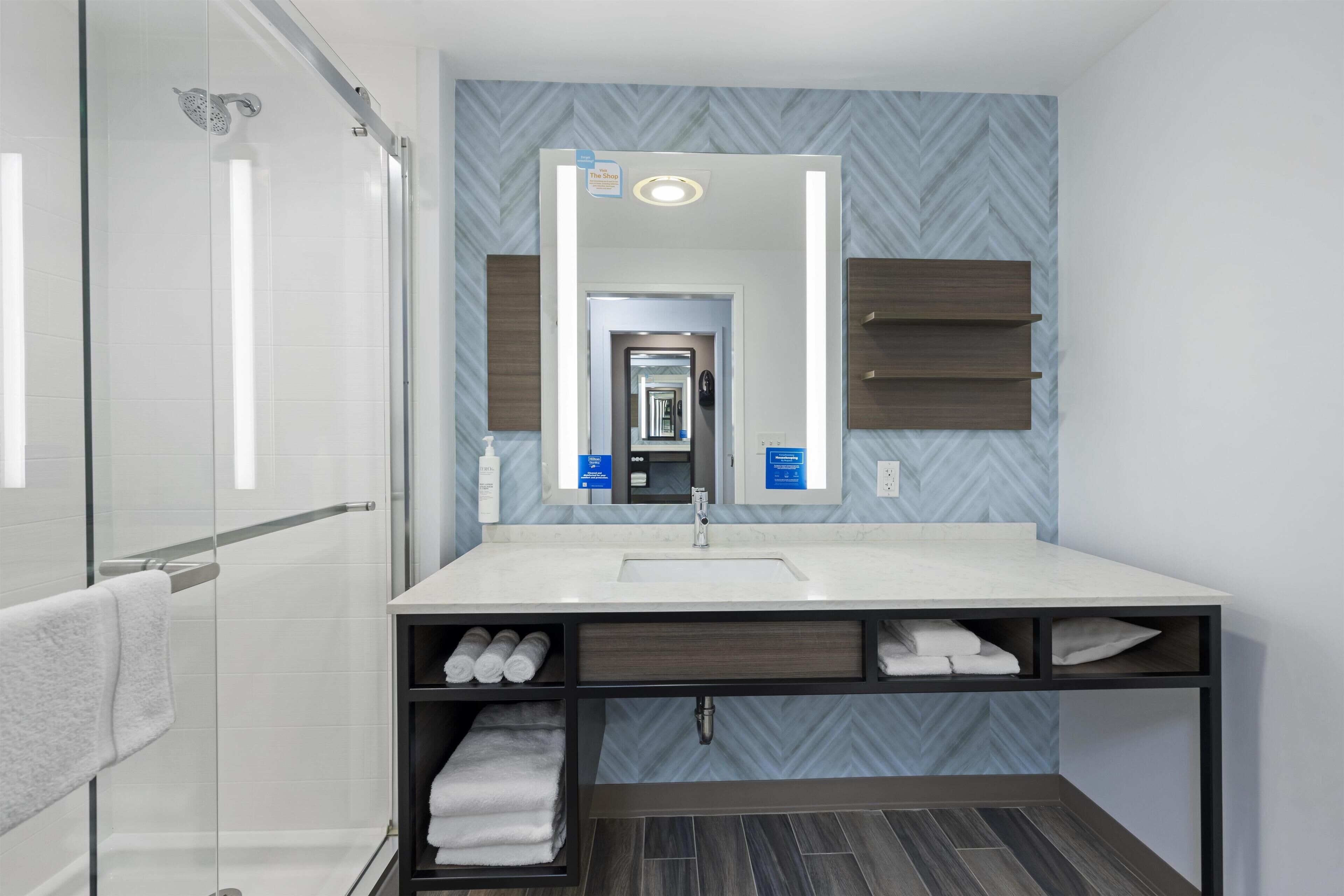 Een moderne badkamer met een douchecabine van glas, een grote spiegel, een wit wastafel en een badmeubel met een donkerhouten afwerking.