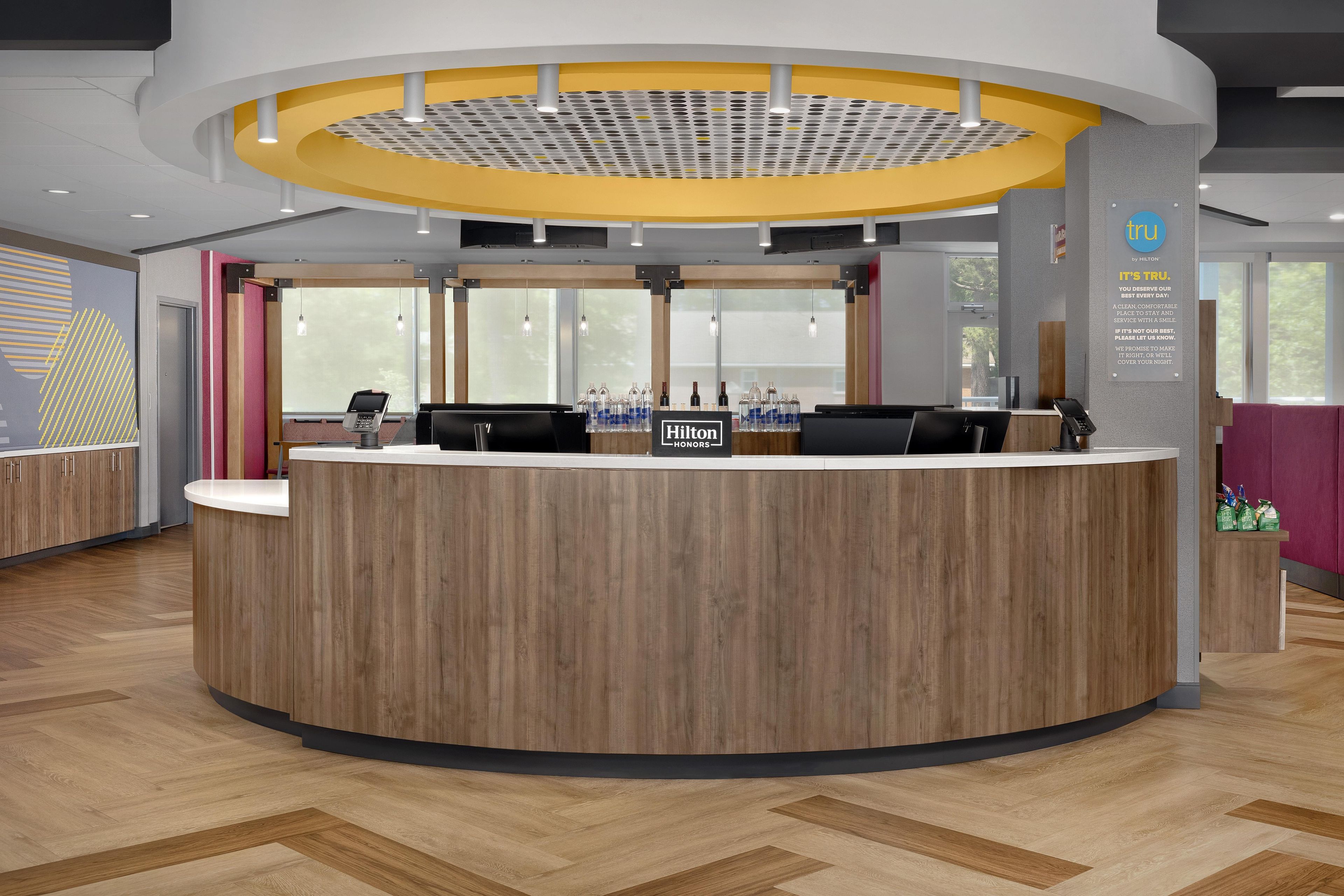 A modern hotel reception area with a wooden reception desk, a digital information kiosk, and a view of the outdoors through large windows.