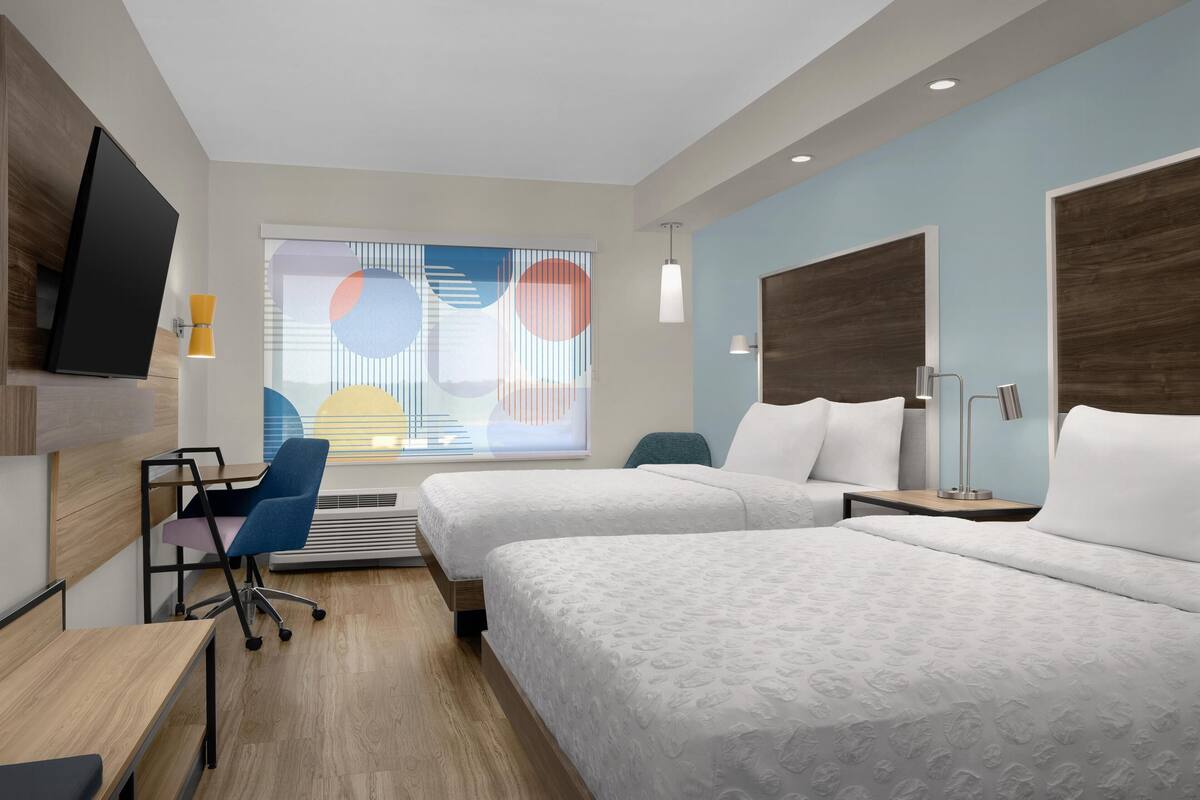A hotel room with two beds, a desk, a chair, a TV, and a window with blinds.