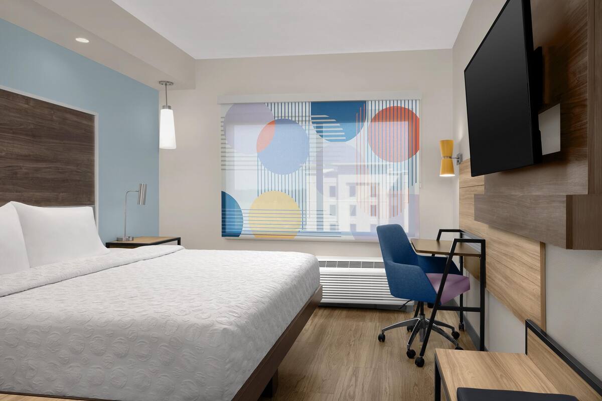 A modern hotel room with a large bed, a desk, a chair, a TV, and a window with a colorful abstract design.