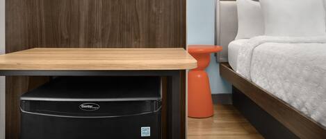 Een hotelkamer met een houten tafel, een zwarte Danby-koelkast, een oranje kruk, een bed met wit beddengoed en een wandlamp.