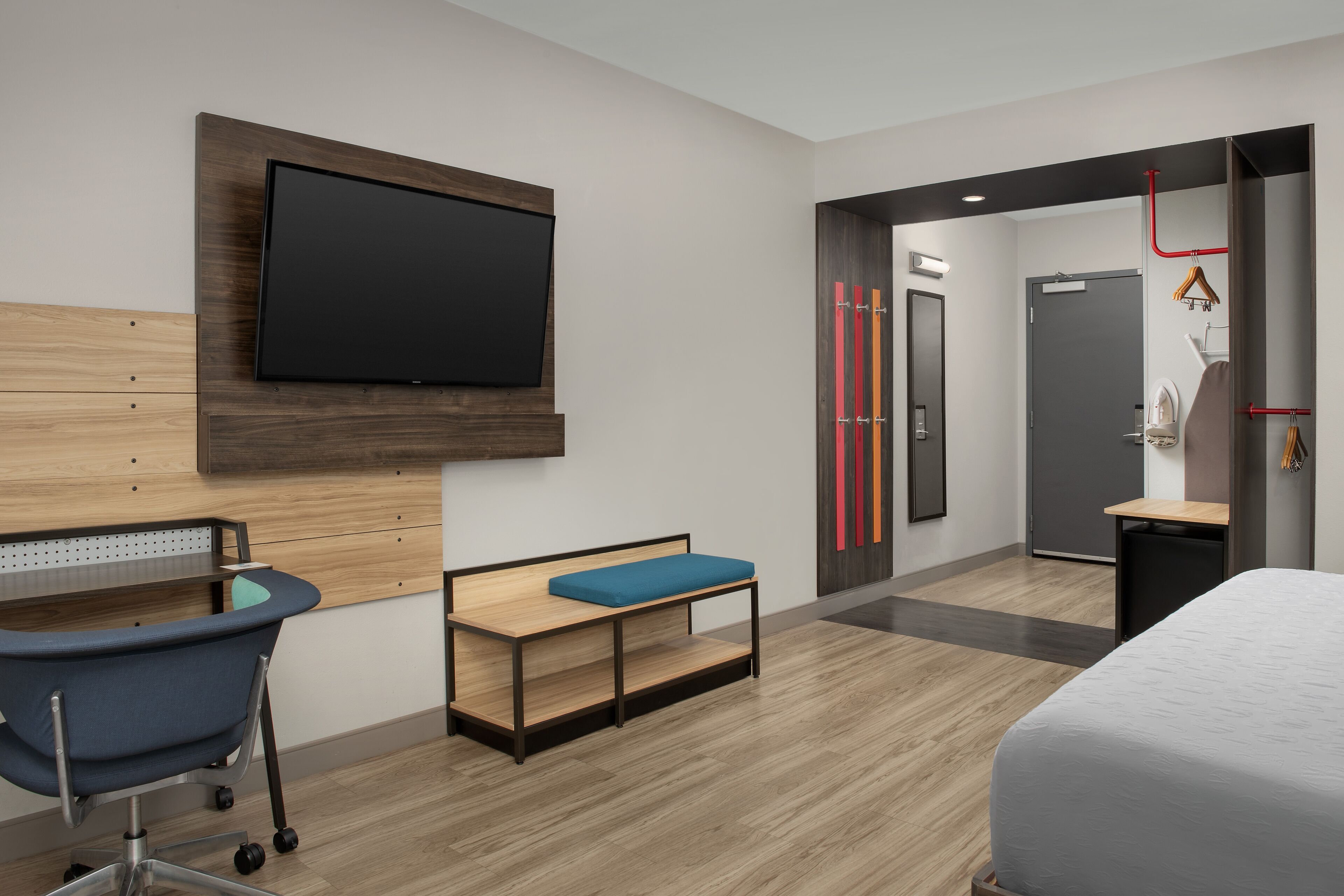 A modern hotel room with a bed, a desk with a chair, a television mounted on the wall, and a small seating area.
