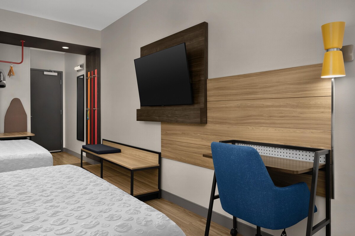 A hotel room with a bed, a desk, a chair, a TV mounted on the wall, and a lamp.