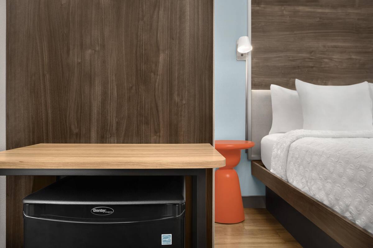 A hotel room with a wooden table, a black Danby refrigerator, an orange stool, a bed with white bedding, and a wall-mounted light.