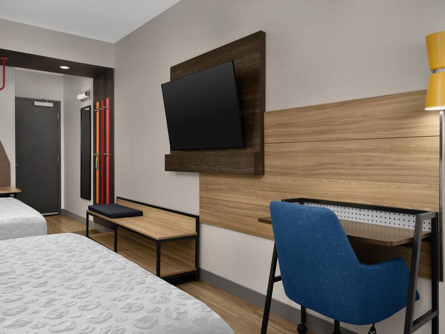 A hotel room with a bed, a desk, a chair, a TV mounted on the wall, and a lamp.
