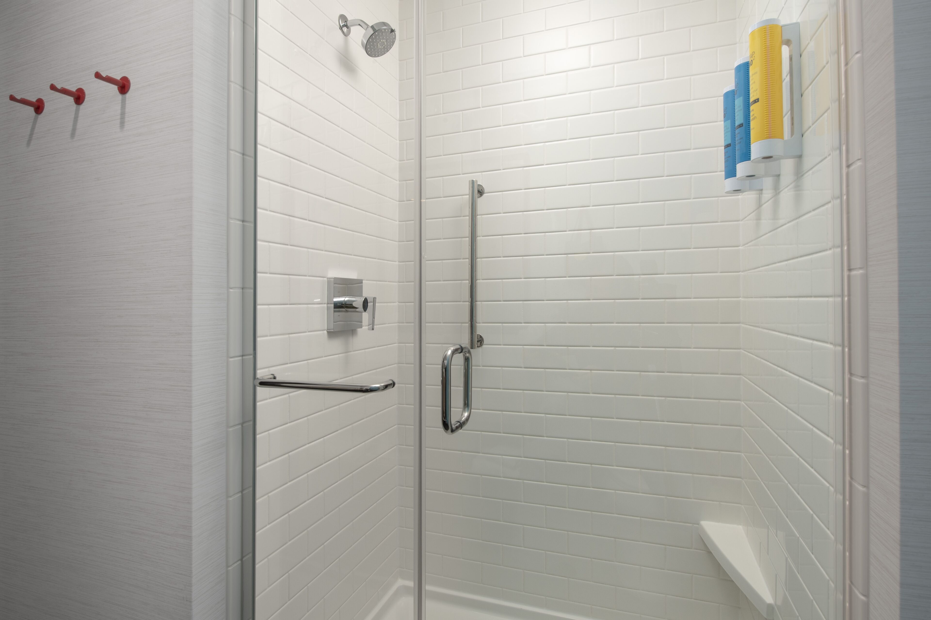 Una ducha moderna con paredes de azulejos blancos, puerta de cristal y repisa con artículos de aseo.