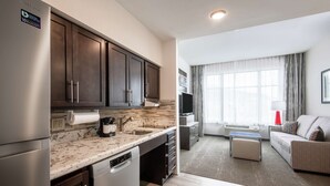 A modern kitchen with dark cabinets, a marble countertop, and a built-in dishwasher. There is a dining area with a sofa and a TV.
