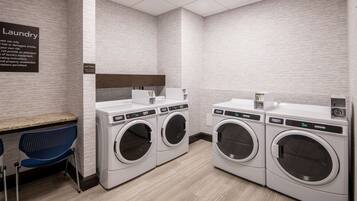 A laundry facility with three front-loading washing machines, a blue chair, and a sign indicating the use of the area.