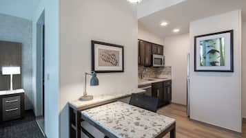 A modern kitchen with a microwave, sink, and refrigerator. There is a dining table with a chair, a desk with a lamp, and framed artwork on the walls.
