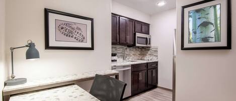 A compact kitchen with dark cabinets, a microwave, and a sink. There is a dining table with a chair and a lamp.