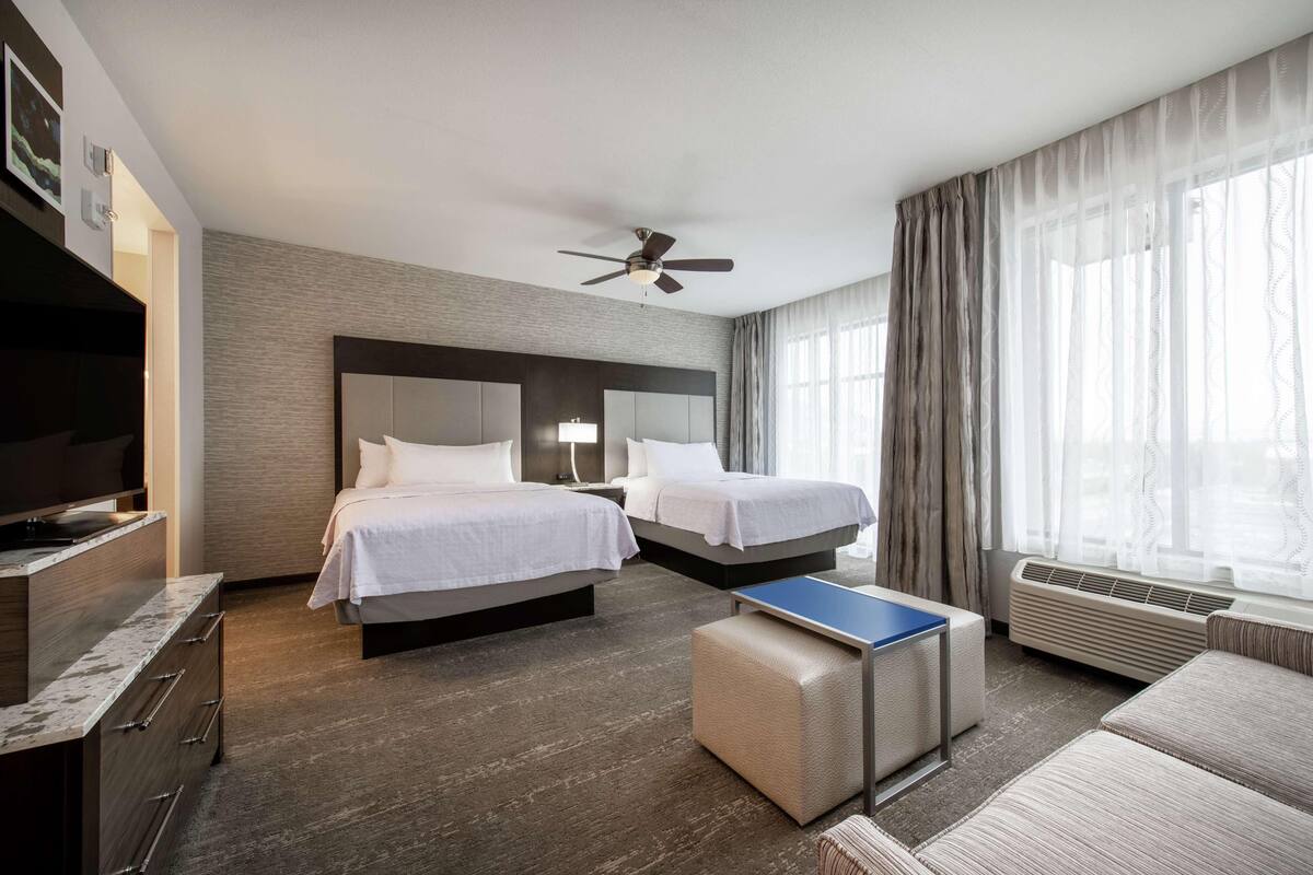Hotellihuone, jossa on kaksi sänkyä, taulutelevisio, kattotuuletin, pieni pöytä ja suuri ikkuna, jossa on verhot.