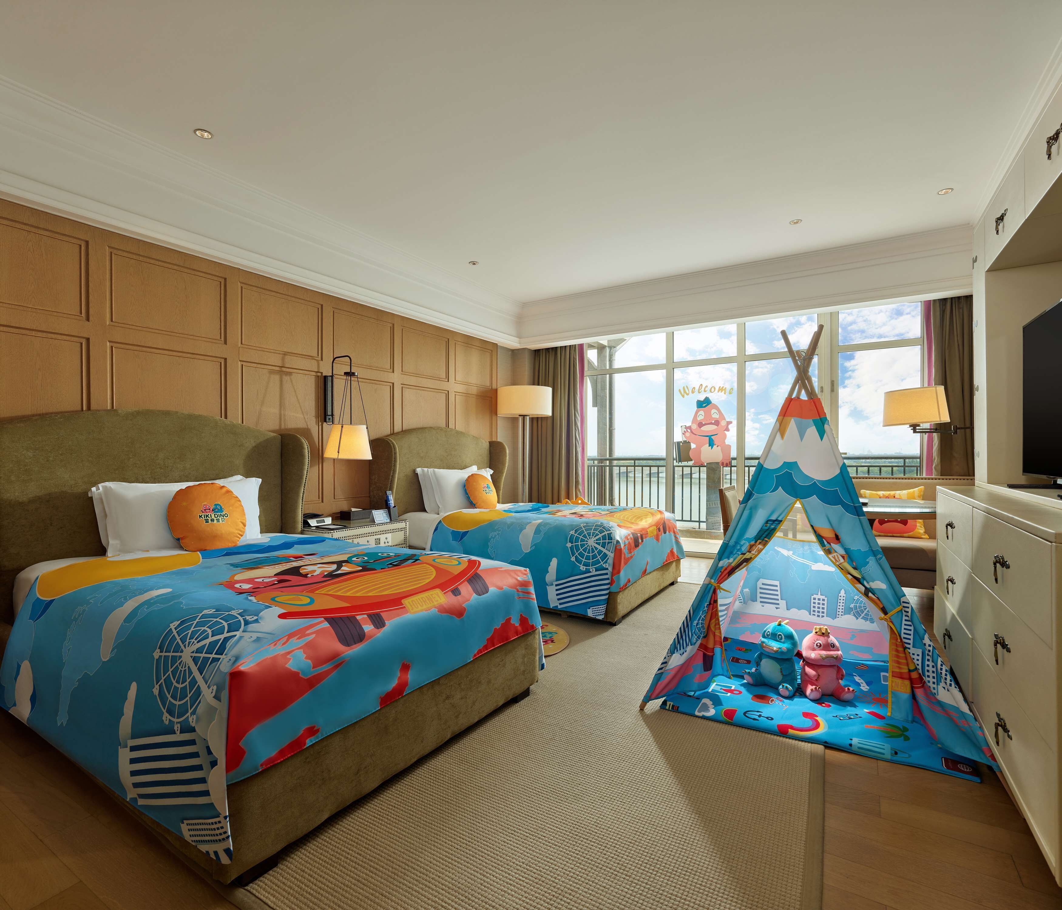 A hotel room with two beds, a large window, a TV, and a children's play area.