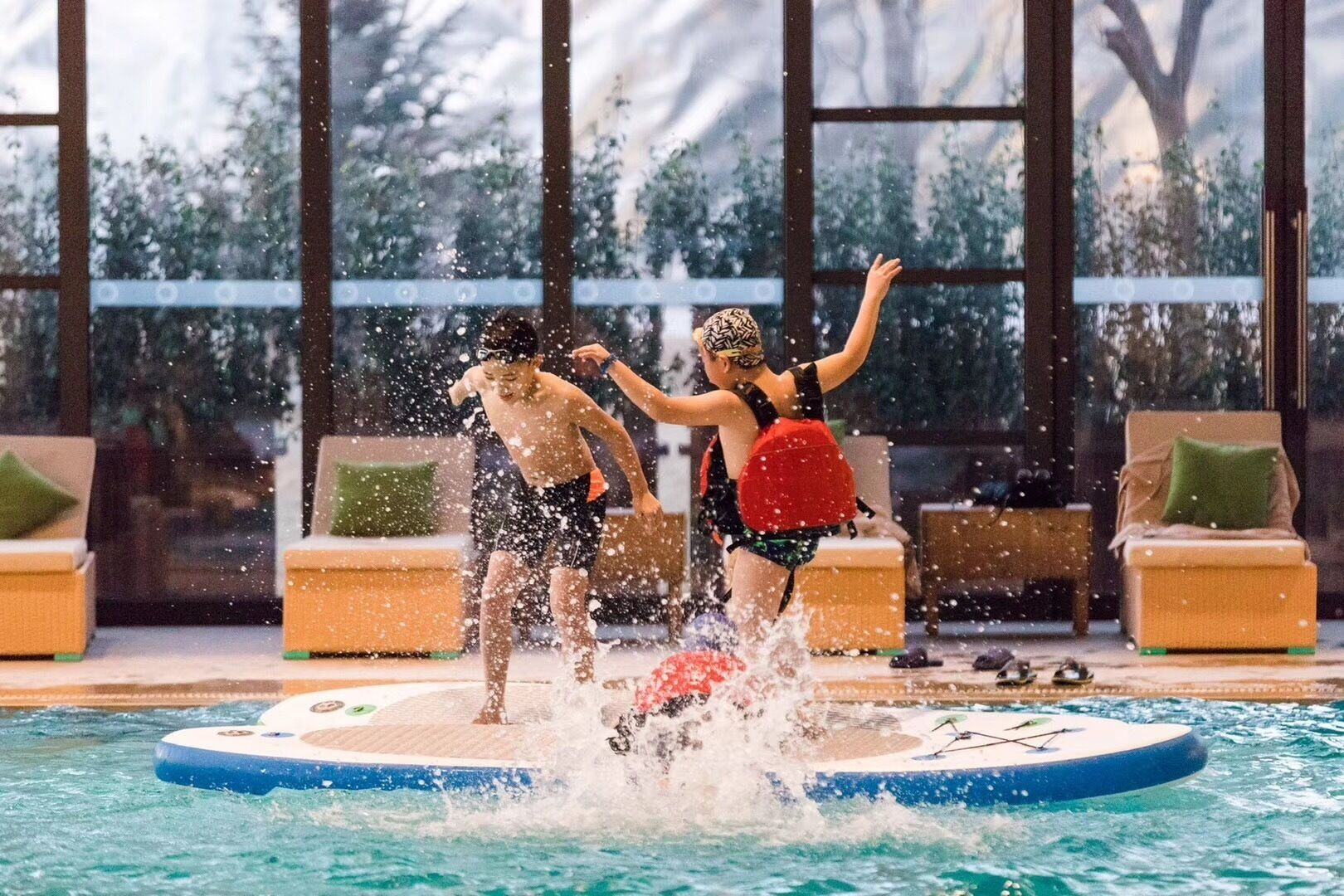 Zona de piscina con una plataforma flotante, sillas de descanso y vistas a árboles a través de amplias ventanas.