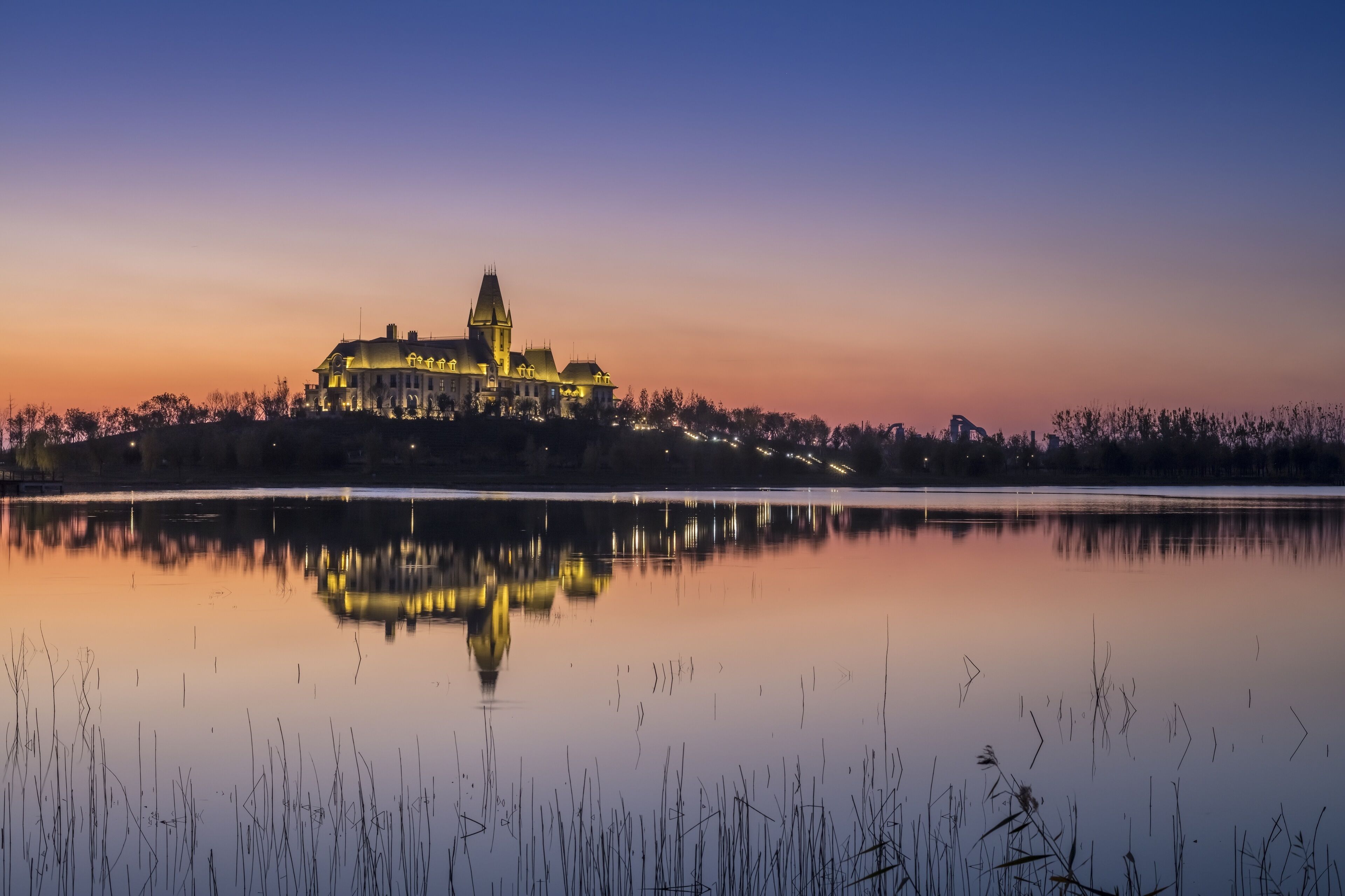 A large, illuminated castle-like building with a prominent tower, surrounded by a calm lake and a forested area.