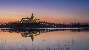 A large, illuminated castle-like building with a prominent tower, surrounded by a calm lake and a forested area.