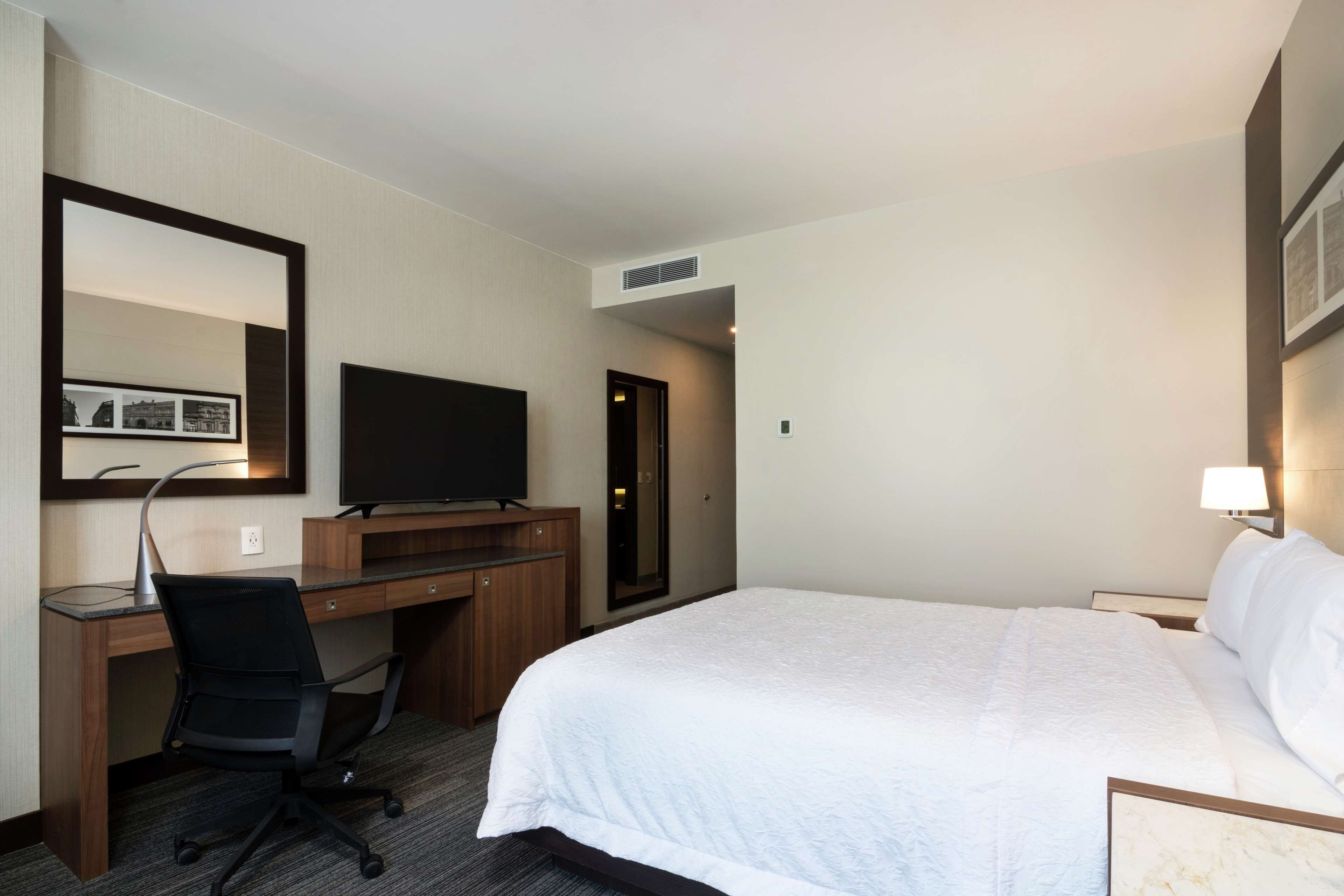 Una habitación de hotel con cama, escritorio, silla, televisor y espejo.