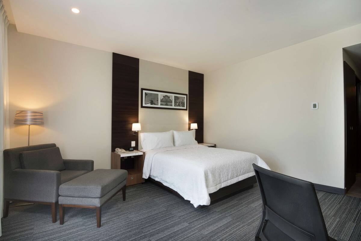 A hotel room with a bed, a chair, a desk, and a lamp.