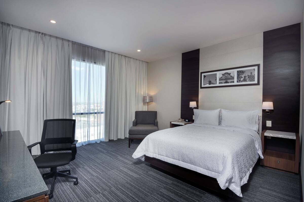 A hotel room with a large bed, a desk, a chair, and a window with curtains. There are wall-mounted lamps and a framed picture above the bed.