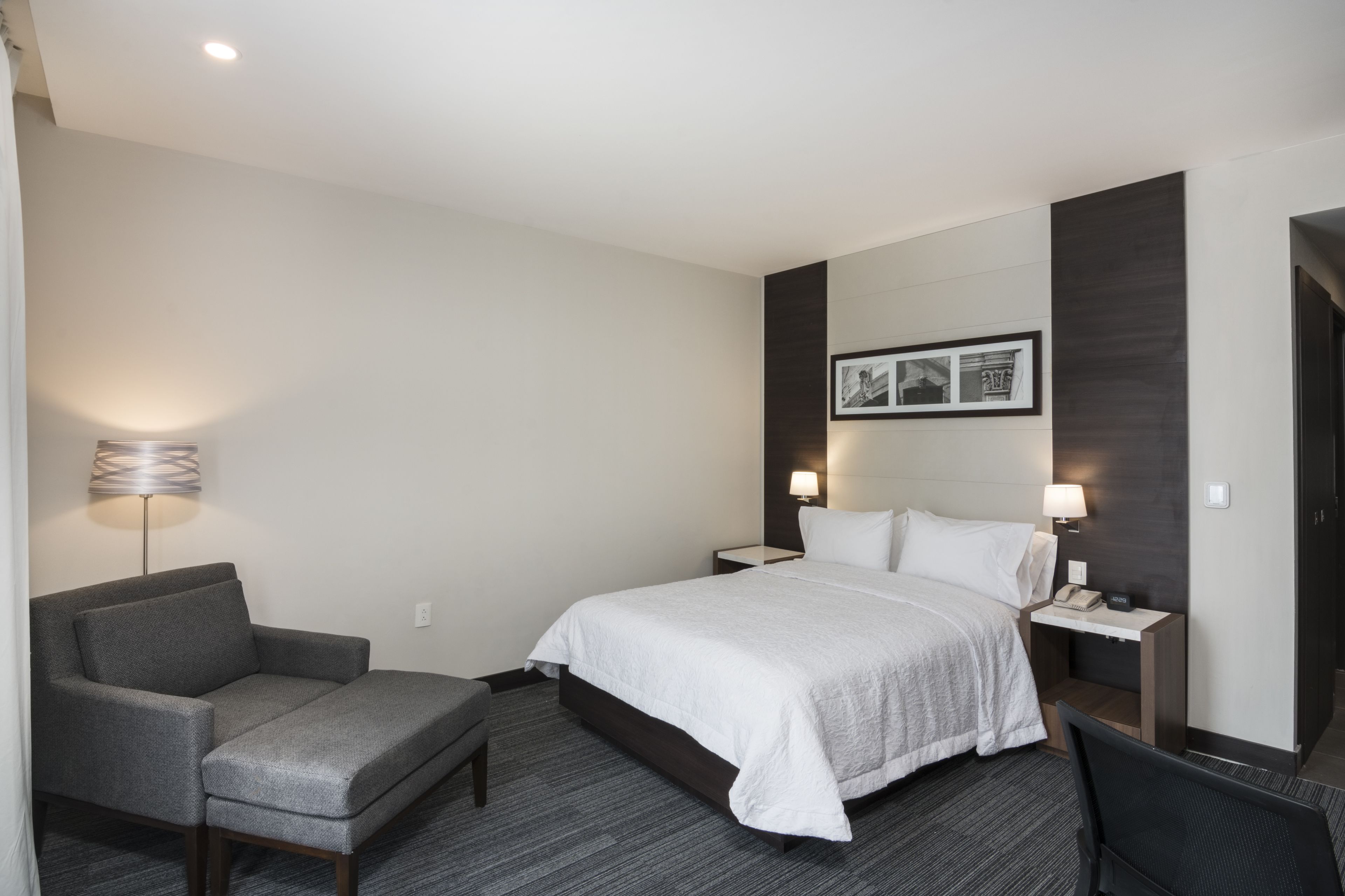 A hotel room with a bed, a chair, a nightstand, a lamp, and a wall-mounted picture.