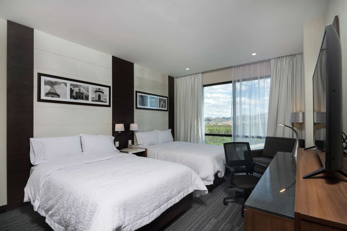 A hotel room with two beds, a desk with a computer, a TV, and large windows with a view.