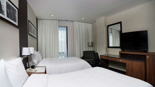A hotel room with two beds, a desk, a chair, a TV, and a large window with curtains.
