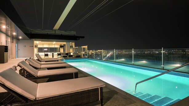 Una piscina infinita en la azotea con sillones de descanso y zona de bar, con vistas a la ciudad por la noche.