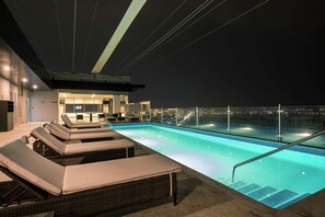 A rooftop infinity pool with lounge chairs and a bar area, offering a city view at night.