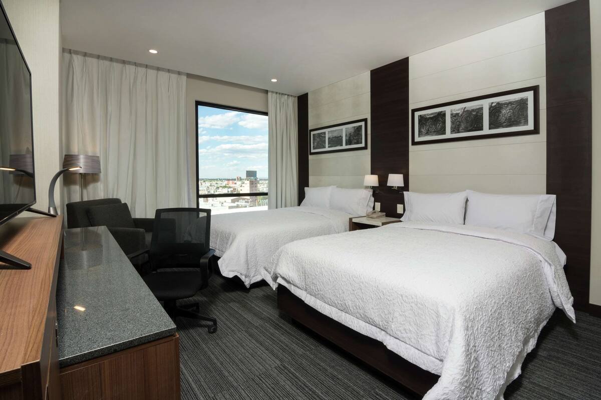 A hotel room with two beds, a desk, a chair, and a view of the city.
