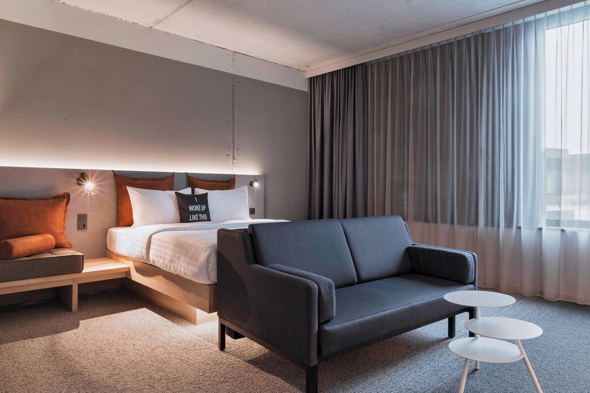 Een moderne hotelkamer met een bed, een bank, een klein tafeltje en een groot raam met gordijnen.