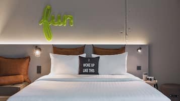 A neatly made bed with a pillow that reads 'I woke up like this,' a neon sign spelling 'fun,' and a wall-mounted light fixture.