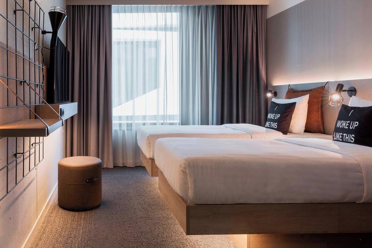 Een hotelkamer met twee bedden, een rond poefje, een groot raam met gordijnen en wandlampen.