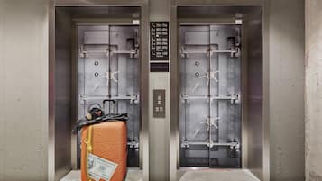 Two elevator doors with a luggage cart containing a suitcase and a handbag, and a sign listing room numbers.