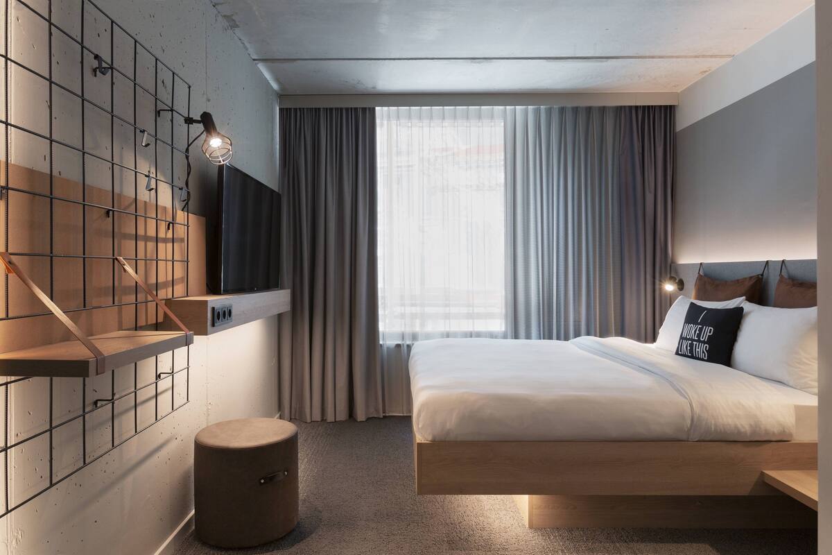 Een moderne hotelkamer met een bed, een wandgemonteerde televisie, een klein krukje en een raam met gordijnen.
