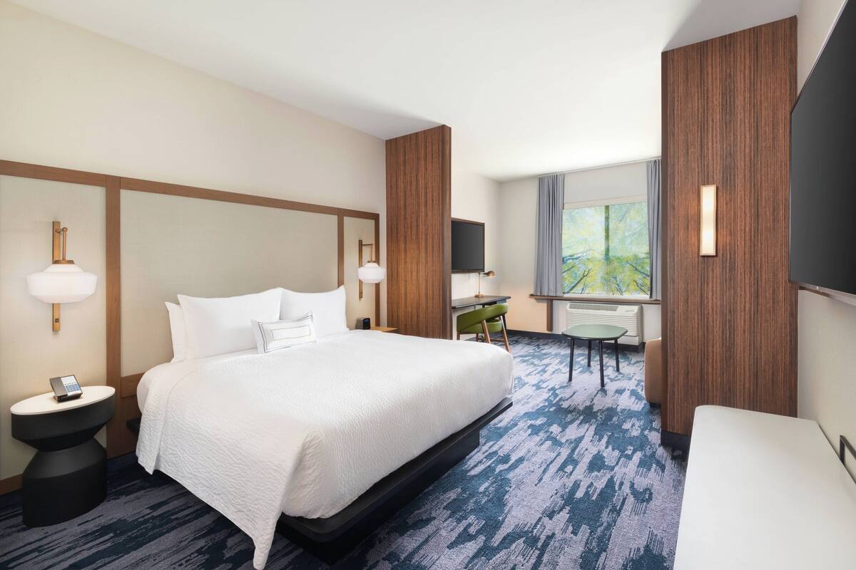 A hotel room with a large bed, a desk, a chair, a TV, and a window with a view of trees.