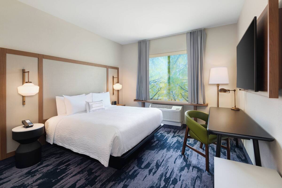 A hotel room with a large bed, bedside tables, a desk, a chair, and a window with a view of trees.