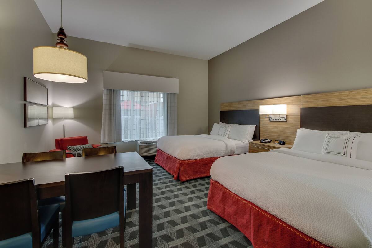 Une chambre d’hôtel avec deux lits, une table à manger, des chaises, une lampe et une fenêtre avec des rideaux.