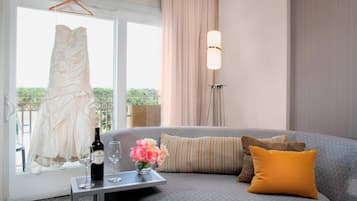 Ein Zimmer mit grauem Sofa, einem Tisch mit Flasche und Gläsern, einem hängenden Kleid und einem Fenster mit Vorhängen.