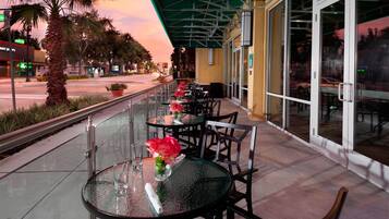 Une terrasse de restaurant avec des tables en plein air, des palmiers et une vue sur la rue au coucher du soleil.