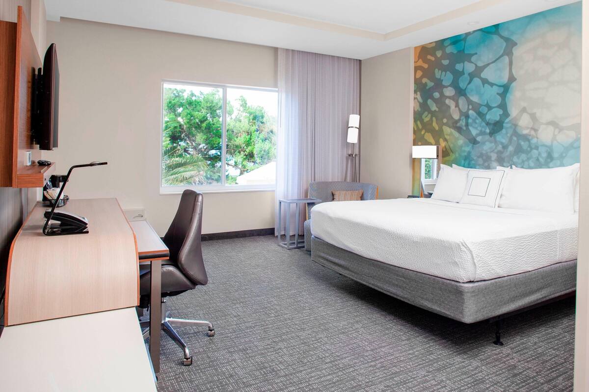 A hotel room with a bed, desk, chair, and a large window with a view of greenery.