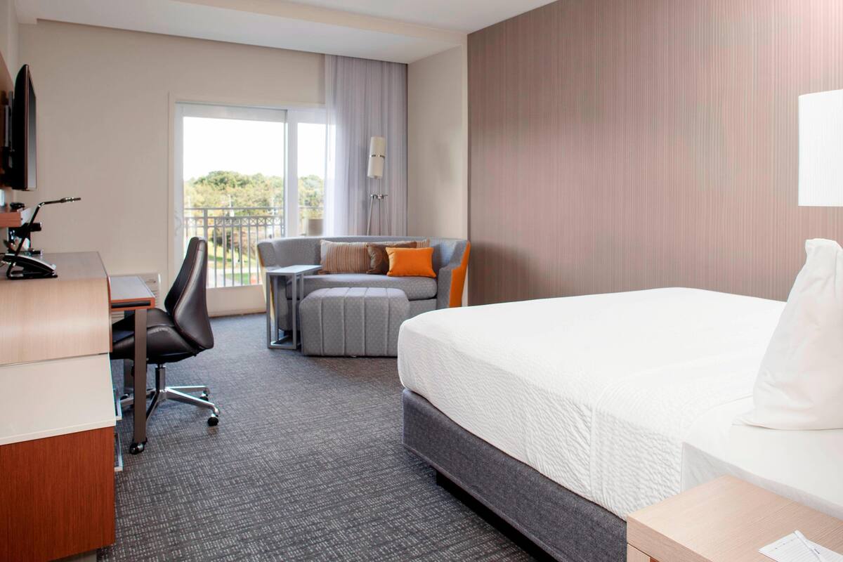 A hotel room with a bed, a desk, a chair, a sofa, and a balcony with a view.