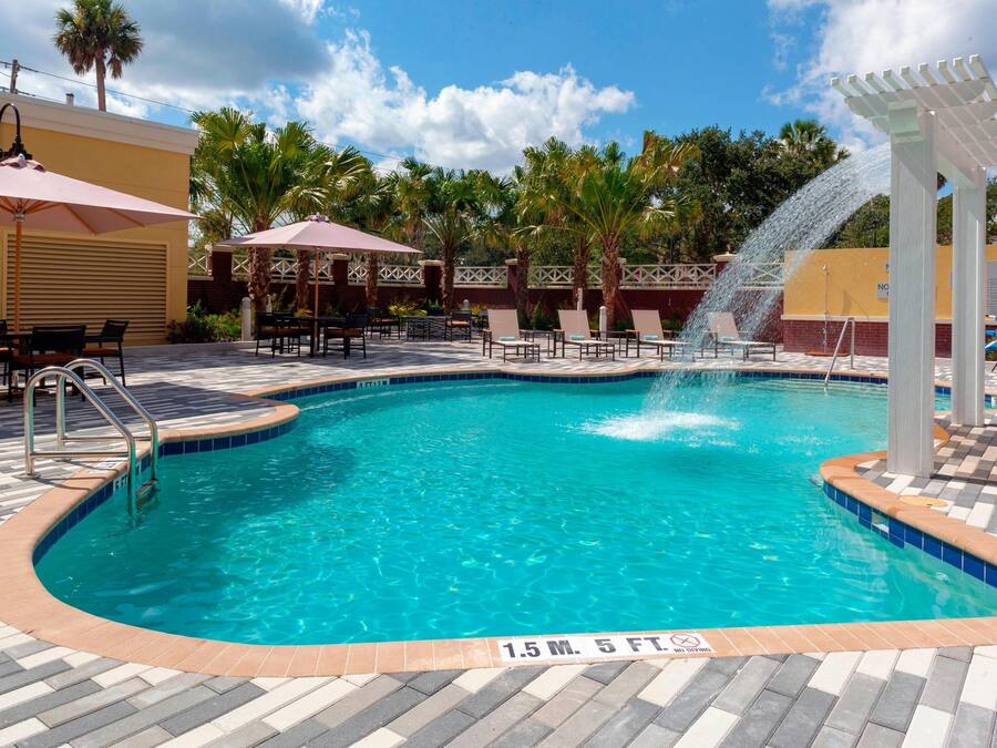 A swimming pool with a water fountain, surrounded by lounge chairs and umbrellas, and palm trees in the background.