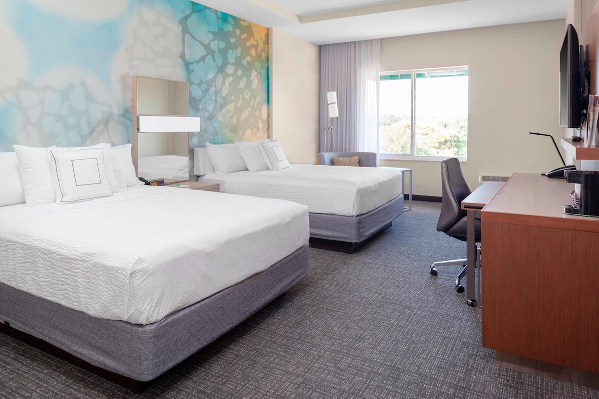 A hotel room with two beds, a desk, a chair, a TV, and a window with a view.