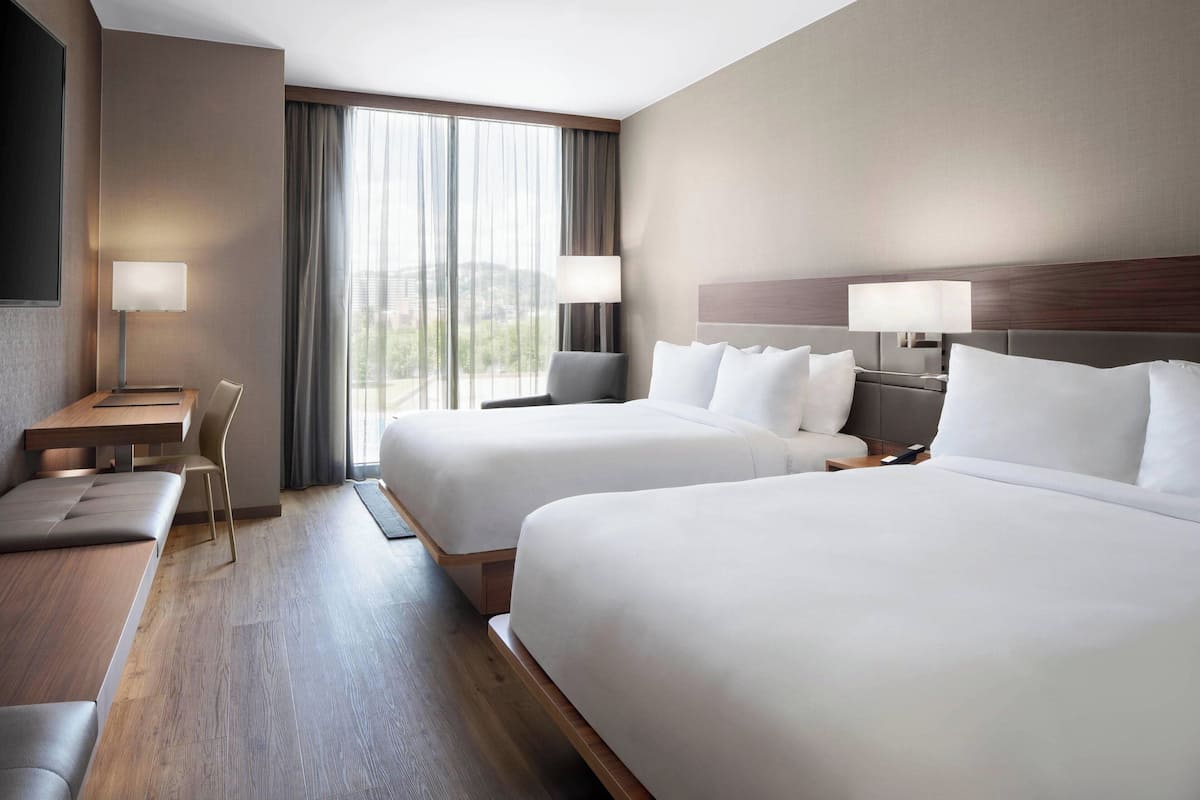A hotel room with two beds, a desk, a chair, and a large window with curtains.
