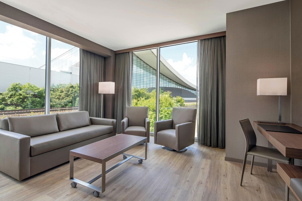 A modern hotel room with a sofa, two armchairs, a desk, and a large window offering a view of greenery and a building.