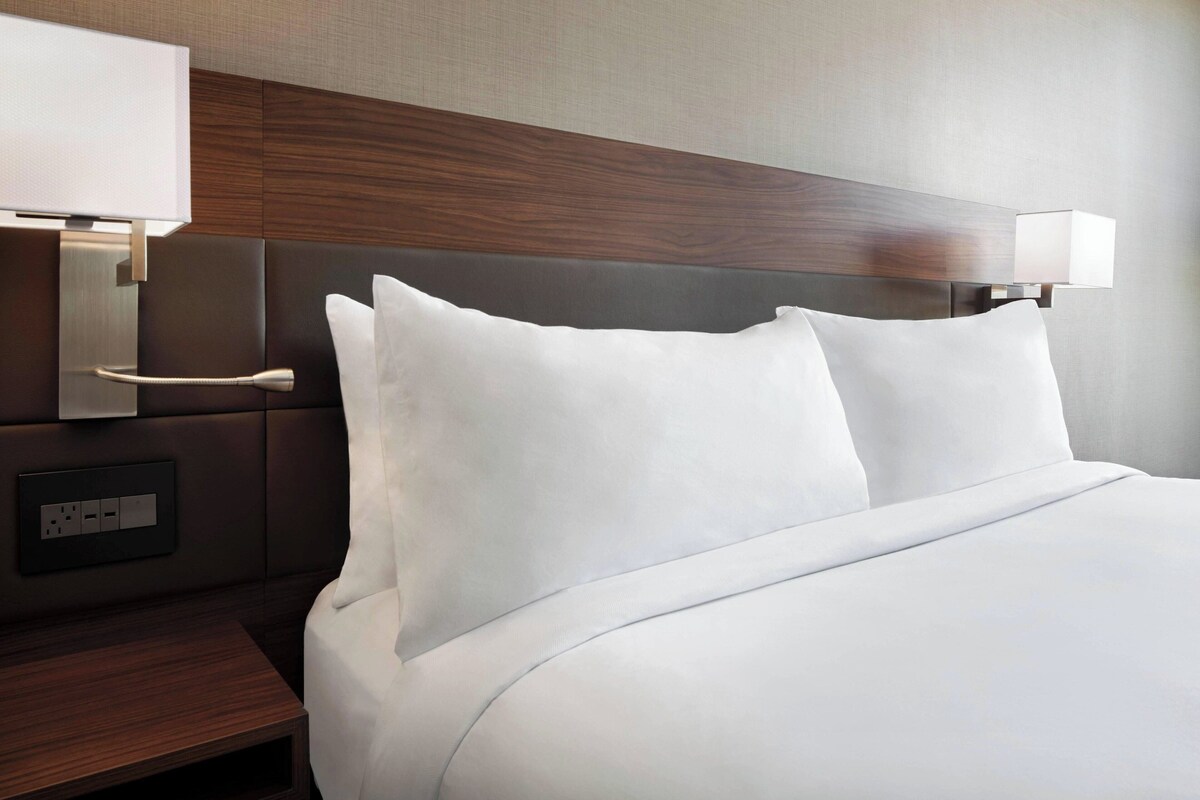 A hotel room with a neatly made bed, a bedside table with a lamp, and a wall-mounted light fixture.