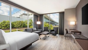 A modern hotel room with a large bed, a sofa, two armchairs, a small table, and a desk. The room has large windows offering a view of a building and greenery. - AC Hotel Pittsburgh Downtown (Pittsburgh)
