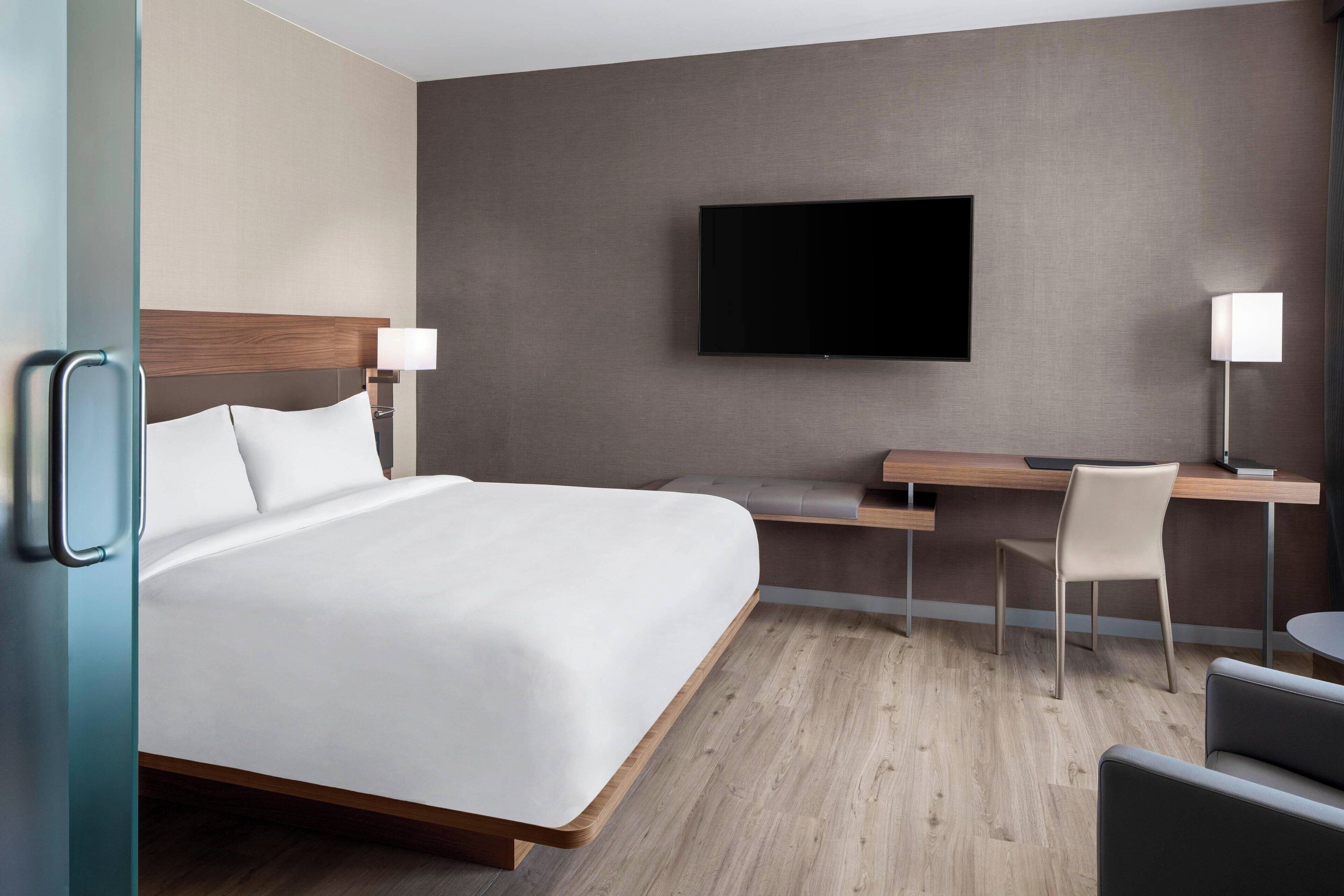 Ein Hotelzimmer mit einem großen Bett, einem Schreibtisch mit Stuhl, einem Flachbildfernseher und einer Lampe.